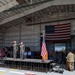 A formal military ceremony is taking place inside a large aircraft hangar. Two officers in flight suits are standing on a raised stage saluting, while a large crowd of military personnel and civilians observe. An American flag is prominently displayed both hanging on the hangar wall and on a flagpole near the podium, suggesting this is likely a change of command or similar official ceremony.
