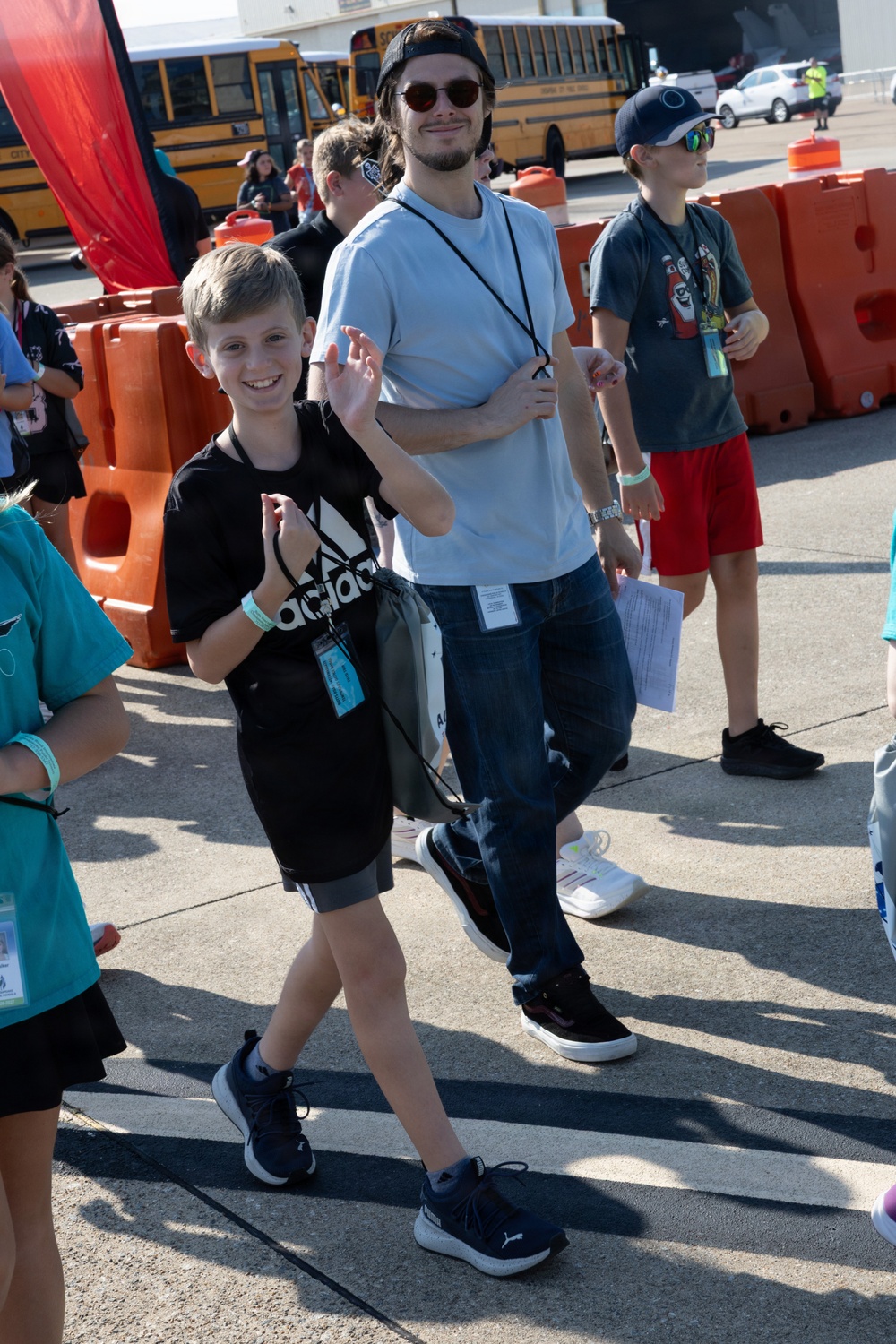 An outdoor event scene showing a group of people, primarily children and young adults, walking in an area marked with orange traffic barriers. A young boy in a black Adidas shirt and a taller young man in a light blue t-shirt appear to be the focal subjects, both wearing lanyards with credentials. Yellow school buses are visible in the background, suggesting a school or youth-organized event.