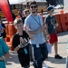 An outdoor event scene showing a group of people, primarily children and young adults, walking in an area marked with orange traffic barriers. A young boy in a black Adidas shirt and a taller young man in a light blue t-shirt appear to be the focal subjects, both wearing lanyards with credentials. Yellow school buses are visible in the background, suggesting a school or youth-organized event.