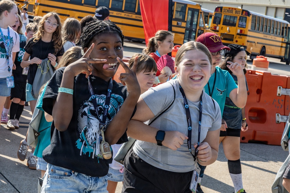 A group of children, likely middle school students, are walking together in an outdoor area near yellow school buses. Two girls in the foreground are posing playfully for the camera, smiling and making hand gestures. The setting appears to be a school field trip or organized event, evidenced by lanyards/badges worn by the students.