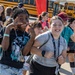 A group of children, likely middle school students, are walking together in an outdoor area near yellow school buses. Two girls in the foreground are posing playfully for the camera, smiling and making hand gestures. The setting appears to be a school field trip or organized event, evidenced by lanyards/badges worn by the students.