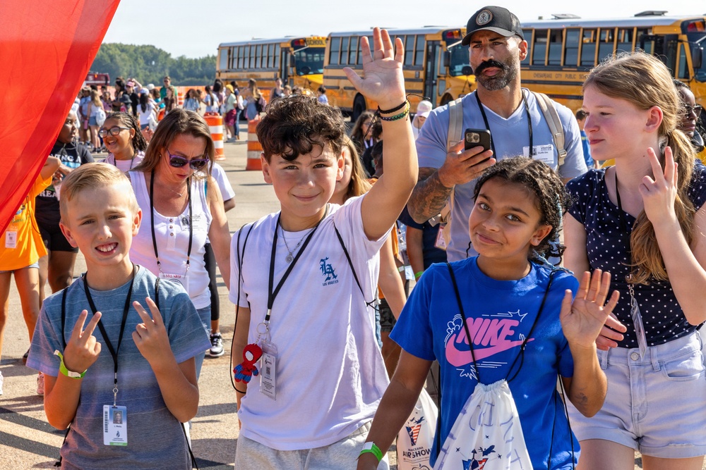 A group of children and adults are gathered outdoors near yellow school buses, appearing to be at an organized field trip or public event, possibly an airshow based on a visible bag logo. Several children are smiling and waving at the camera, wearing lanyards with ID badges. The atmosphere is cheerful and energetic under bright sunny conditions.