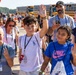 A group of children and adults are gathered outdoors near yellow school buses, appearing to be at an organized field trip or public event, possibly an airshow based on a visible bag logo. Several children are smiling and waving at the camera, wearing lanyards with ID badges. The atmosphere is cheerful and energetic under bright sunny conditions.