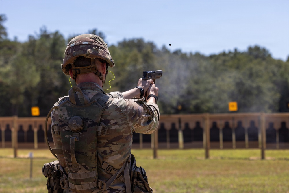 A soldier in full combat gear is firing a pistol at a military shooting range. The image captures a spent casing being ejected from the firearm mid-shot, with multiple silhouette targets visible downrange. The setting appears to be an outdoor military training facility with a tree line in the background.