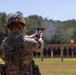 A soldier in full combat gear is firing a pistol at a military shooting range. The image captures a spent casing being ejected from the firearm mid-shot, with multiple silhouette targets visible downrange. The setting appears to be an outdoor military training facility with a tree line in the background.