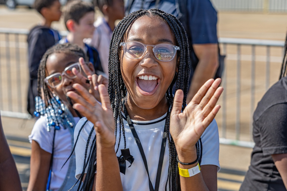 A young girl with braided hair and clear-framed glasses is captured mid-expression, clapping her hands with an open-mouthed smile showing great excitement or enthusiasm. She is wearing a white striped shirt, a black lanyard, and a yellow wristband. The outdoor setting appears to be a public event with a metal barrier and other children visible in the background.