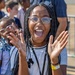 A young girl with braided hair and clear-framed glasses is captured mid-expression, clapping her hands with an open-mouthed smile showing great excitement or enthusiasm. She is wearing a white striped shirt, a black lanyard, and a yellow wristband. The outdoor setting appears to be a public event with a metal barrier and other children visible in the background.