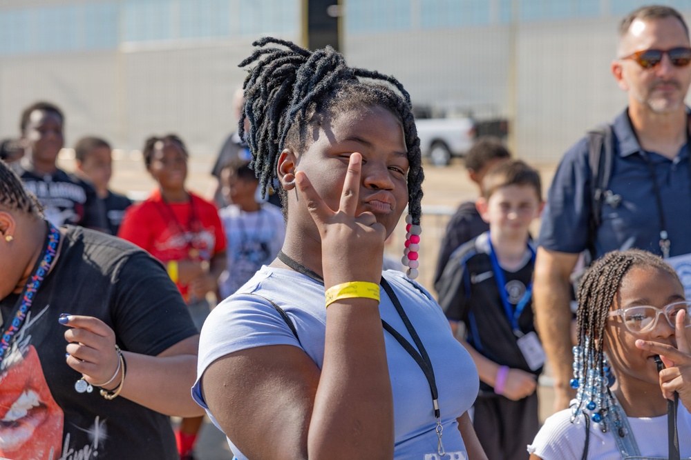 A group of children and youth are gathered outdoors, likely at an organized event or field trip, as evidenced by the lanyards and wristbands visible on several participants. The central figure, a young girl with loc hairstyle, is posing playfully for the camera with a peace sign gesture near her eye. An adult male supervisor wearing sunglasses is visible in the background.