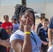 A group of children and youth are gathered outdoors, likely at an organized event or field trip, as evidenced by the lanyards and wristbands visible on several participants. The central figure, a young girl with loc hairstyle, is posing playfully for the camera with a peace sign gesture near her eye. An adult male supervisor wearing sunglasses is visible in the background.
