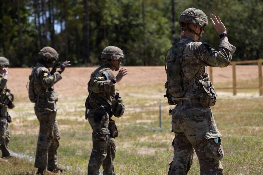 Multiple military personnel in camouflage uniforms and helmets are standing in a line on a training range, all with their hands raised. The setting appears to be an outdoor military training facility with a dirt range area, wooden fence, and tree line in the background. The raised hands gesture suggests a safety halt or range command during a live-fire or tactical training exercise.