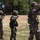 2026 US ARMY BEST SQUAD COMPETITION: Pistol Match Multiple military personnel in camouflage uniforms and helmets are standing in a line on a training range, all with their hands raised. The setting appears to be an outdoor military training facility with a dirt range area, wooden fence, and tree line in the background. The raised hands gesture suggests a safety halt or range command during a live-fire or tactical training exercise.