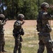 Multiple military personnel in camouflage uniforms and helmets are standing in a line on a training range, all with their hands raised. The setting appears to be an outdoor military training facility with a dirt range area, wooden fence, and tree line in the background. The raised hands gesture suggests a safety halt or range command during a live-fire or tactical training exercise.