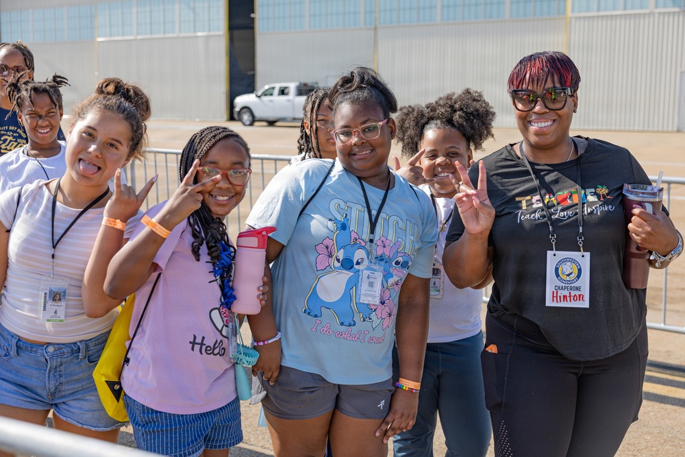 A group of young students and a chaperone pose together outdoors, smiling and making peace signs and playful gestures for the camera. The adult on the right is wearing a badge identifying her as 'Chaperone Hinton' and a shirt reading 'Teach, Love, Inspire.' The setting appears to be a field trip at what looks like an airfield or industrial facility.