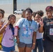 A group of young students and a chaperone pose together outdoors, smiling and making peace signs and playful gestures for the camera. The adult on the right is wearing a badge identifying her as 'Chaperone Hinton' and a shirt reading 'Teach, Love, Inspire.' The setting appears to be a field trip at what looks like an airfield or industrial facility.