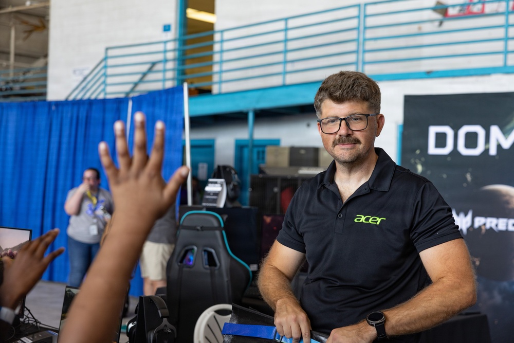 A man wearing an Acer-branded polo shirt is staffing what appears to be an Acer/Predator gaming booth at a technology or gaming expo. He is interacting with a visitor whose raised hand is visible in the foreground. The booth features gaming chairs, monitors, and Acer Predator branding in the background.