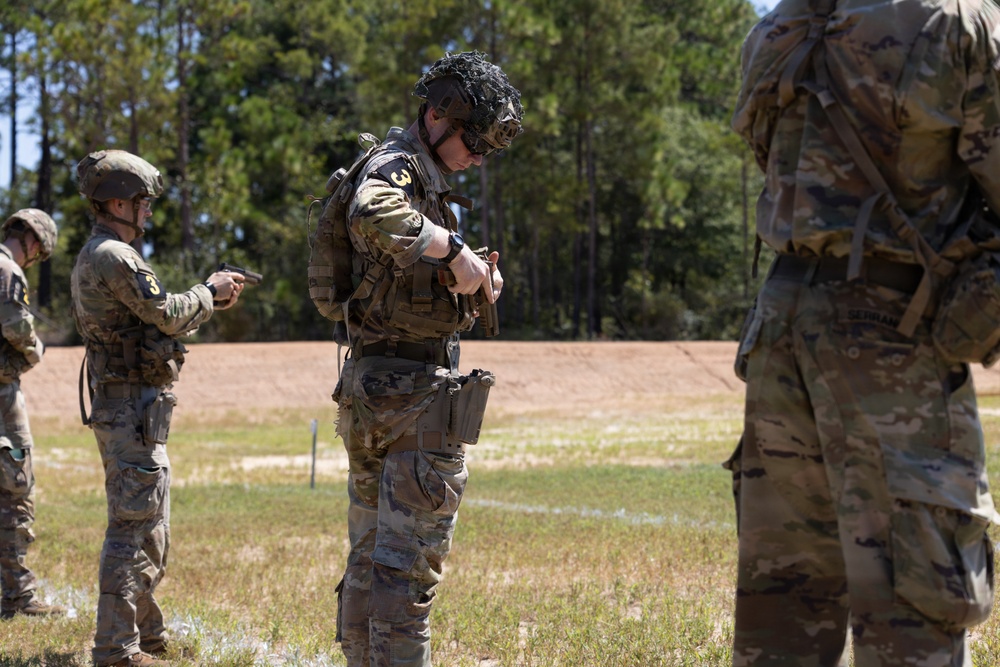 Military personnel in MultiCam/OCP camouflage uniforms are conducting what appears to be a weapons handling or equipment inspection exercise at an outdoor range or training area. At least three soldiers are visible, with the central figure wearing a helmet with netting and tactical gear bearing the number '3'. The setting appears to be a military training facility with a cleared field and tree line in the background.