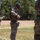 2026 US ARMY BEST SQUAD COMPETITION: Pistol Match Military personnel in MultiCam/OCP camouflage uniforms are conducting what appears to be a weapons handling or equipment inspection exercise at an outdoor range or training area. At least three soldiers are visible, with the central figure wearing a helmet with netting and tactical gear bearing the number '3'. The setting appears to be a military training facility with a cleared field and tree line in the background.