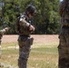 Military personnel in MultiCam/OCP camouflage uniforms are conducting what appears to be a weapons handling or equipment inspection exercise at an outdoor range or training area. At least three soldiers are visible, with the central figure wearing a helmet with netting and tactical gear bearing the number '3'. The setting appears to be a military training facility with a cleared field and tree line in the background.
