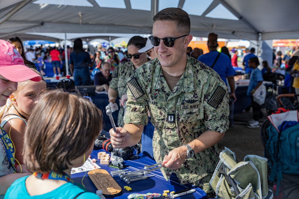 A U.S. Navy service member in NWU Type III camouflage uniform is engaging with children at what appears to be a public outreach or community event. The sailor is leaning over a display table showing tools and equipment to young visitors under a white tent. A second uniformed Navy personnel member is visible in the background.