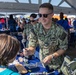 A U.S. Navy service member in NWU Type III camouflage uniform is engaging with children at what appears to be a public outreach or community event. The sailor is leaning over a display table showing tools and equipment to young visitors under a white tent. A second uniformed Navy personnel member is visible in the background.