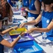 An outdoor community event or science fair where a young volunteer or staff member in a blue t-shirt is assisting a child with a hands-on water activity at a table. Multiple colored plastic bins filled with soapy water are arranged on a white table, suggesting a bubble or cleaning science experiment. Several other participants and volunteers in matching blue shirts are visible in the background.