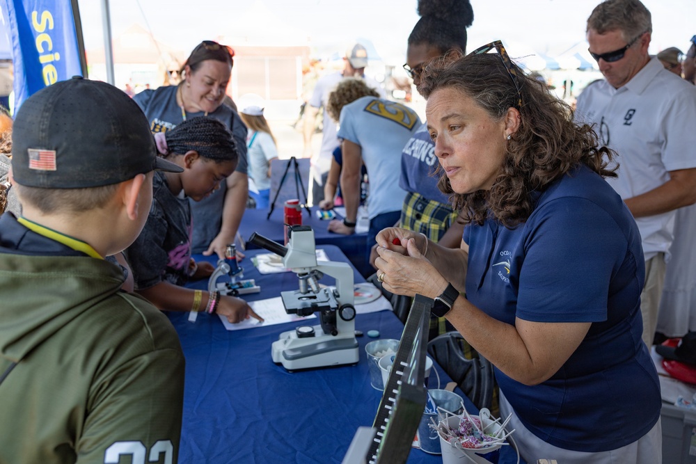 An outdoor science education event where adults in matching blue shirts engage children at a demonstration table. A woman in the foreground appears to be explaining or demonstrating something small to a child wearing a cap with an American flag patch. Microscopes and educational materials are visible on the blue-draped table.