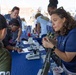 An outdoor science education event where adults in matching blue shirts engage children at a demonstration table. A woman in the foreground appears to be explaining or demonstrating something small to a child wearing a cap with an American flag patch. Microscopes and educational materials are visible on the blue-draped table.