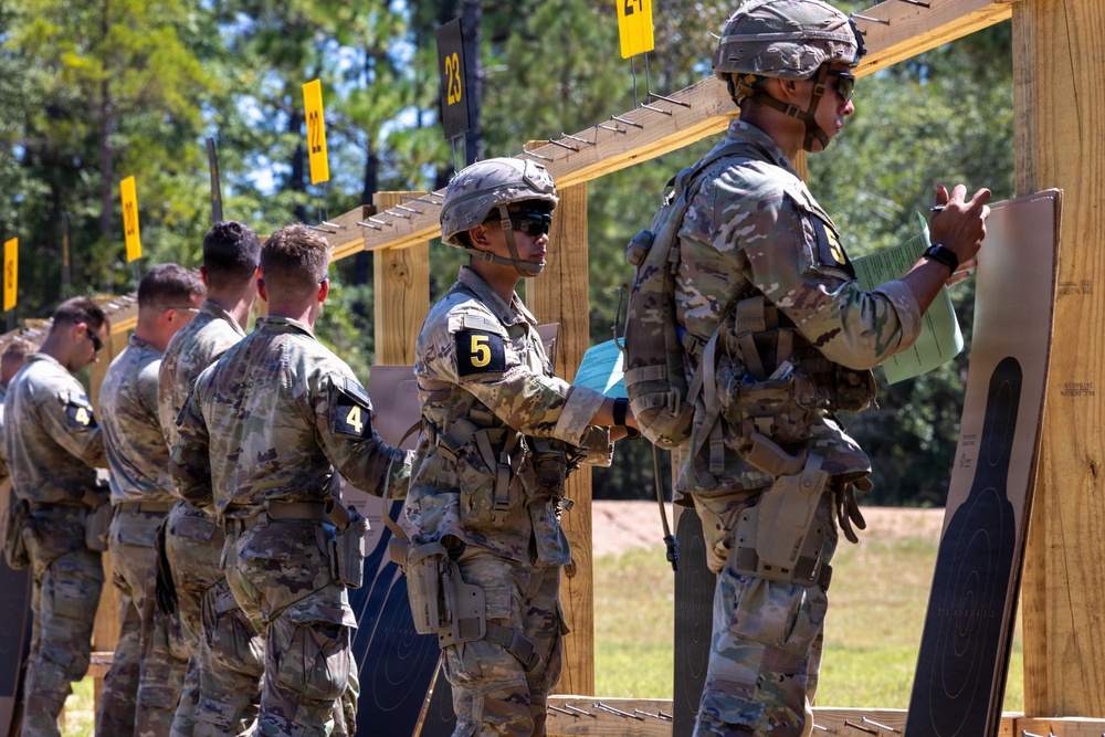 Military personnel in camouflage uniforms are lined up at a shooting range, examining paper targets mounted on a wooden frame structure. Soldiers are numbered with yellow placards (visible numbers include 4 and 5), suggesting a scored competition or qualification event. The setting appears to be an outdoor range with trees visible in the background.