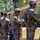 2026 US ARMY BEST SQUAD COMPETITION: Pistol Match Military personnel in camouflage uniforms are lined up at a shooting range, examining paper targets mounted on a wooden frame structure. Soldiers are numbered with yellow placards (visible numbers include 4 and 5), suggesting a scored competition or qualification event. The setting appears to be an outdoor range with trees visible in the background.