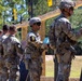 Military personnel in camouflage uniforms are lined up at a shooting range, examining paper targets mounted on a wooden frame structure. Soldiers are numbered with yellow placards (visible numbers include 4 and 5), suggesting a scored competition or qualification event. The setting appears to be an outdoor range with trees visible in the background.