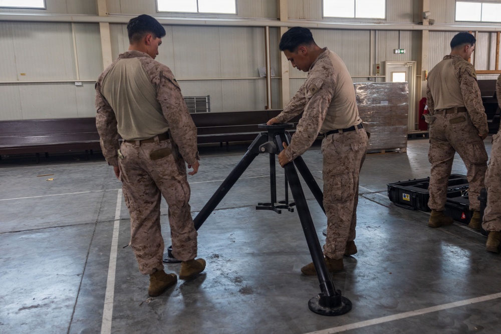 Several military personnel in desert camouflage uniforms are working inside a large indoor facility, assembling or handling what appears to be a tripod-style military equipment stand. Two soldiers in the foreground are focused on manipulating the black metal tripod structure, while a third soldier is visible in the background near additional equipment. The setting appears to be a warehouse or hangar-type building.