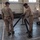 Several military personnel in desert camouflage uniforms are working inside a large indoor facility, assembling or handling what appears to be a tripod-style military equipment stand. Two soldiers in the foreground are focused on manipulating the black metal tripod structure, while a third soldier is visible in the background near additional equipment. The setting appears to be a warehouse or hangar-type building.