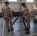Several military personnel in desert camouflage uniforms are working inside a large indoor facility, assembling or handling what appears to be a tripod-style military equipment stand. Two soldiers in the foreground are focused on manipulating the black metal tripod structure, while a third soldier is visible in the background near additional equipment. The setting appears to be a warehouse or hangar-type building.