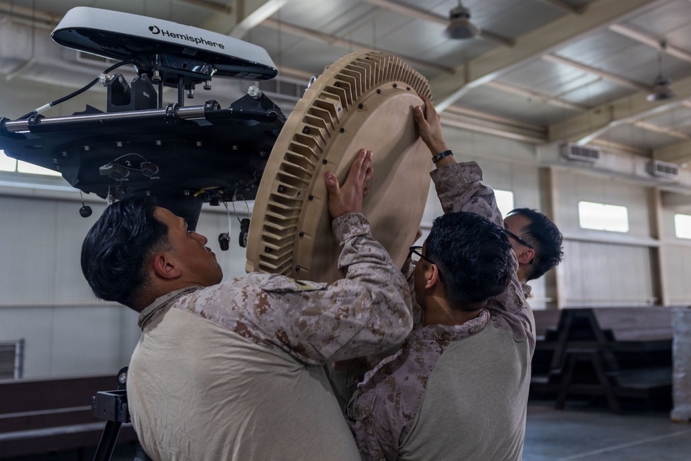 Three military personnel in desert camouflage uniforms are collaboratively installing or assembling a large circular antenna or radar component onto a mounting system inside a hangar or warehouse facility. The equipment includes a visible 'Hemisphere' branded GPS/navigation unit mounted above the circular dish component. The scene depicts a technical military equipment assembly or maintenance operation.