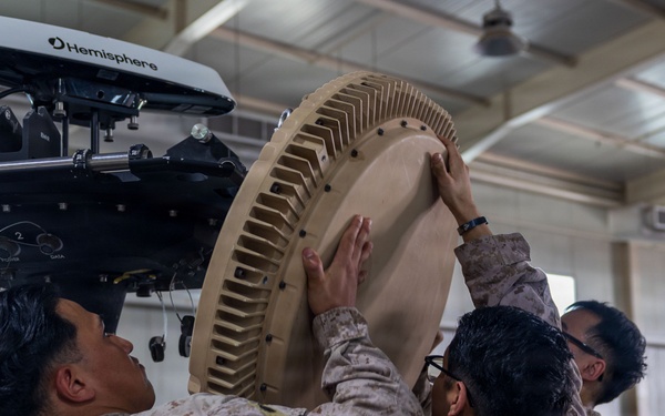 Three military personnel in desert camouflage uniforms are collaboratively installing or assembling a large circular antenna or radar component onto a mounting system inside a hangar or warehouse facility. The equipment includes a visible 'Hemisphere' branded GPS/navigation unit mounted above the circular dish component. The scene depicts a technical military equipment assembly or maintenance operation.