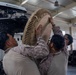 Three military personnel in desert camouflage uniforms are collaboratively installing or assembling a large circular antenna or radar component onto a mounting system inside a hangar or warehouse facility. The equipment includes a visible 'Hemisphere' branded GPS/navigation unit mounted above the circular dish component. The scene depicts a technical military equipment assembly or maintenance operation.