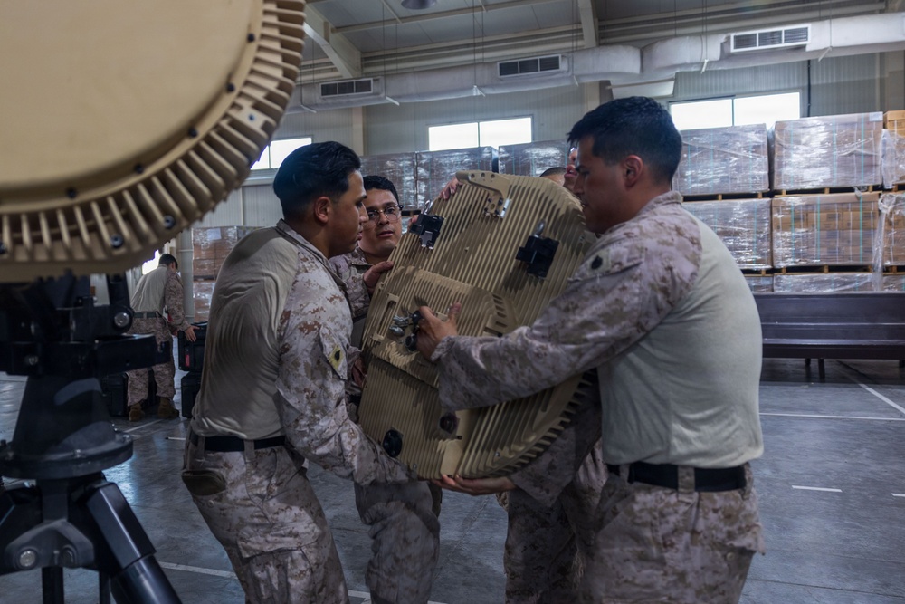 Several military personnel in desert camouflage uniforms are handling a large tan-colored electronic or communications equipment component inside what appears to be a warehouse or hangar facility. The soldiers appear to be assembling, installing, or repositioning the equipment, which has a ribbed/finned housing consistent with radar or communications hardware. Additional palletized cargo wrapped in plastic is visible in the background.