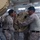 Several military personnel in desert camouflage uniforms are handling a large tan-colored electronic or communications equipment component inside what appears to be a warehouse or hangar facility. The soldiers appear to be assembling, installing, or repositioning the equipment, which has a ribbed/finned housing consistent with radar or communications hardware. Additional palletized cargo wrapped in plastic is visible in the background.