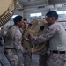 Several military personnel in desert camouflage uniforms are handling a large tan-colored electronic or communications equipment component inside what appears to be a warehouse or hangar facility. The soldiers appear to be assembling, installing, or repositioning the equipment, which has a ribbed/finned housing consistent with radar or communications hardware. Additional palletized cargo wrapped in plastic is visible in the background.
