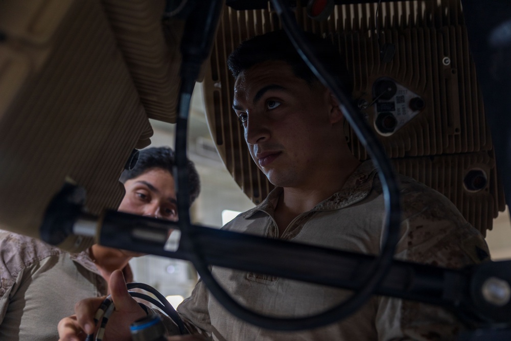Two military personnel in desert camouflage uniforms are closely examining or working on a piece of equipment. The image is taken from a low angle, showing the individuals focused on what appears to be a technical or mechanical component with cables and metal framing. The lighting is dramatic and low, creating strong shadows across their faces.