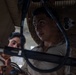 Two military personnel in desert camouflage uniforms are closely examining or working on a piece of equipment. The image is taken from a low angle, showing the individuals focused on what appears to be a technical or mechanical component with cables and metal framing. The lighting is dramatic and low, creating strong shadows across their faces.