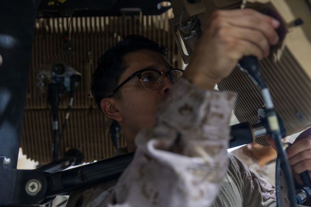 A person wearing glasses and what appears to be military camouflage uniform is working on mechanical or technical equipment, viewed from a low angle. They appear to be performing maintenance or installation work on components mounted overhead or at an elevated position. The setting appears to be inside a vehicle or mechanical workspace with tools visible in the background.