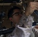 A person wearing glasses and what appears to be military camouflage uniform is working on mechanical or technical equipment, viewed from a low angle. They appear to be performing maintenance or installation work on components mounted overhead or at an elevated position. The setting appears to be inside a vehicle or mechanical workspace with tools visible in the background.