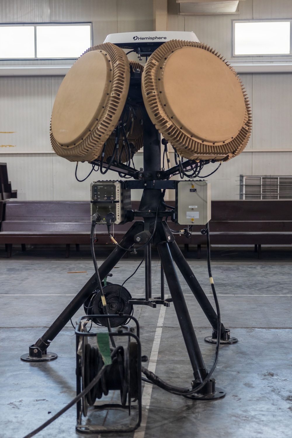 The image shows a military or tactical radar/antenna system mounted on a black tripod stand inside an indoor facility, likely a hangar or training hall. The system features two large circular phased-array antenna dishes in a tan/desert color scheme, with a 'Hemisphere' branded GPS/navigation unit mounted on top. Various control boxes, cables, and a cable reel are visible as part of the integrated system.