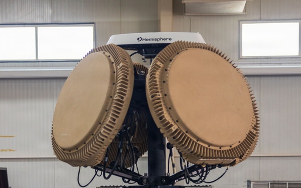 The image shows a military or tactical radar/antenna system mounted on a black tripod stand inside an indoor facility, likely a hangar or training hall. The system features two large circular phased-array antenna dishes in a tan/desert color scheme, with a 'Hemisphere' branded GPS/navigation unit mounted on top. Various control boxes, cables, and a cable reel are visible as part of the integrated system.