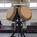 The image shows a military or tactical radar/antenna system mounted on a black tripod stand inside an indoor facility, likely a hangar or training hall. The system features two large circular phased-array antenna dishes in a tan/desert color scheme, with a 'Hemisphere' branded GPS/navigation unit mounted on top. Various control boxes, cables, and a cable reel are visible as part of the integrated system.