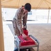 A military service member in desert camouflage (MARPAT) uniform is fueling or servicing a portable generator using red fuel canisters. The scene appears to be at a forward operating base or military installation, indicated by the modular buildings and paved courtyard visible in the background. At least two red fuel canisters are present, one marked '11TH MEU' indicating Marine Expeditionary Unit affiliation.