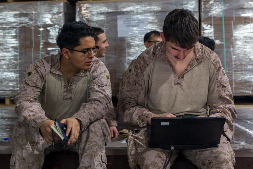 Two military personnel in desert camouflage uniforms are seated together, with one operating a laptop computer while the other observes and holds a small notebook. Additional personnel are visible in the background near what appears to be palletized cargo wrapped in plastic shrink wrap. The setting appears to be a warehouse or logistics staging area.
