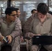 Two military personnel in desert camouflage uniforms are seated together, with one operating a laptop computer while the other observes and holds a small notebook. Additional personnel are visible in the background near what appears to be palletized cargo wrapped in plastic shrink wrap. The setting appears to be a warehouse or logistics staging area.