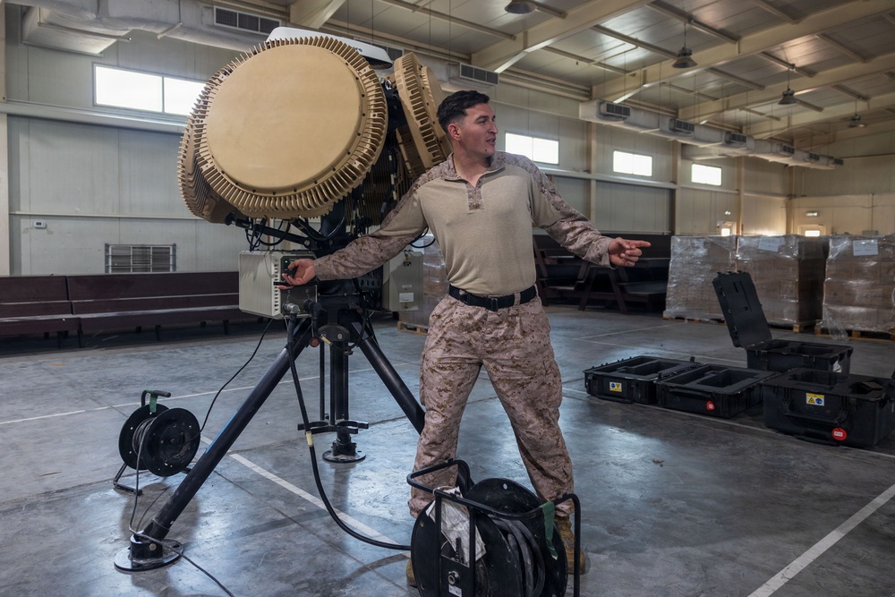 A military service member in desert camouflage uniform stands beside a large tripod-mounted radar or directed-energy equipment inside a warehouse or hangar facility. The individual appears to be gesturing or explaining something, possibly conducting a demonstration or briefing. The setting includes military equipment cases and cable reels on a concrete floor.